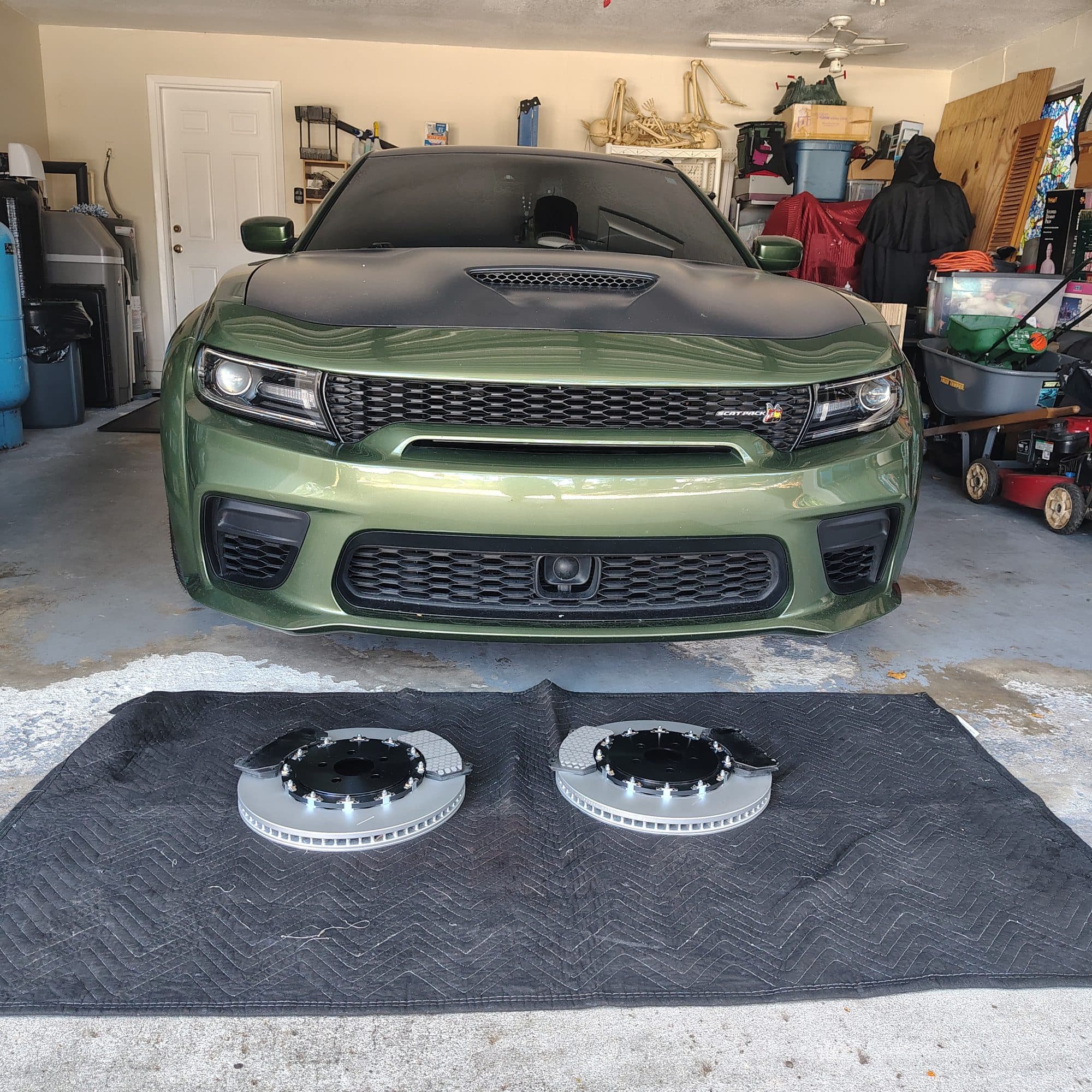 A green Dodge Charger Scat Pack parked in a garage with two large two-piece brake rotors laid out on a mat in front of it, ready to go on