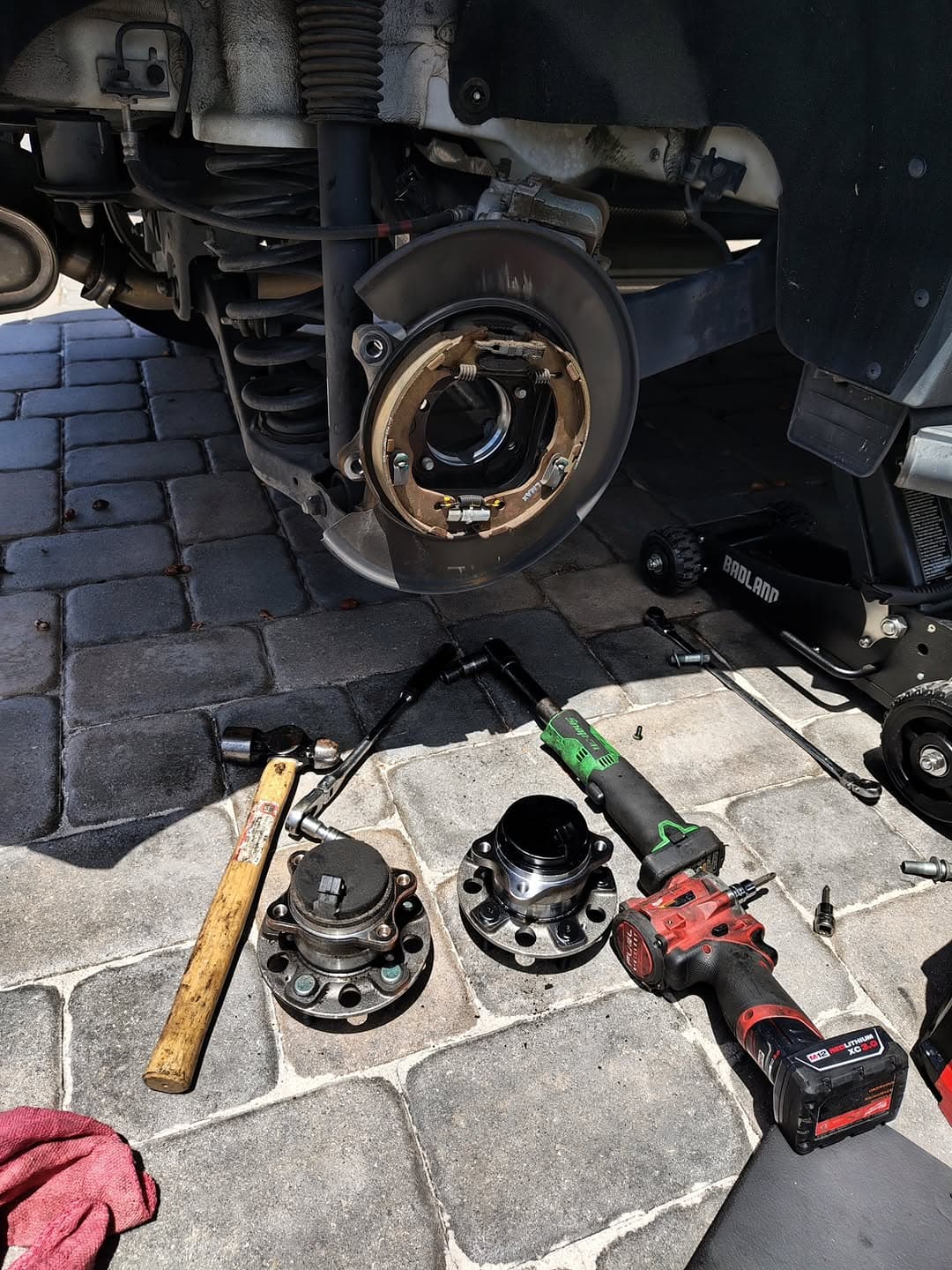 A rear wheel bearing job on a driveway: the hub carrier stripped down on the car, with the old and new bearing assemblies and an impact gun laid out on the pavers