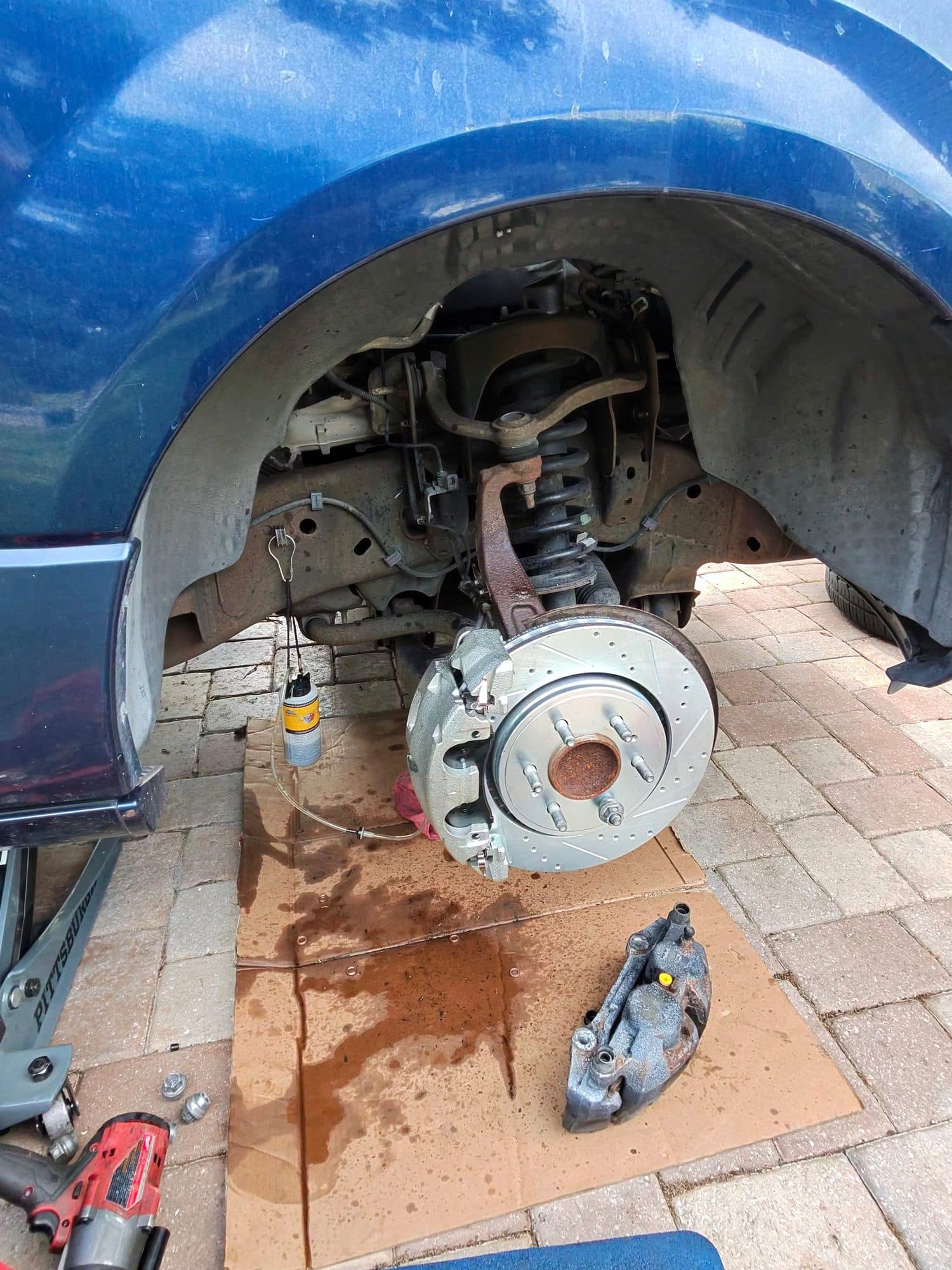A front brake job on a blue truck in a paver driveway: a new drilled and slotted rotor and caliper fitted, the old caliper bracket and an impact gun laid out on cardboard below