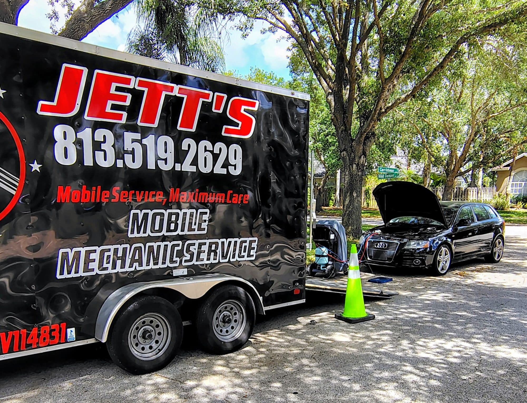 The Jett's Mobile Mechanic Service trailer parked on a shady residential street, its phone number down the side, with a black Audi wagon behind it, hood open and equipment set out on the ground beside the fender