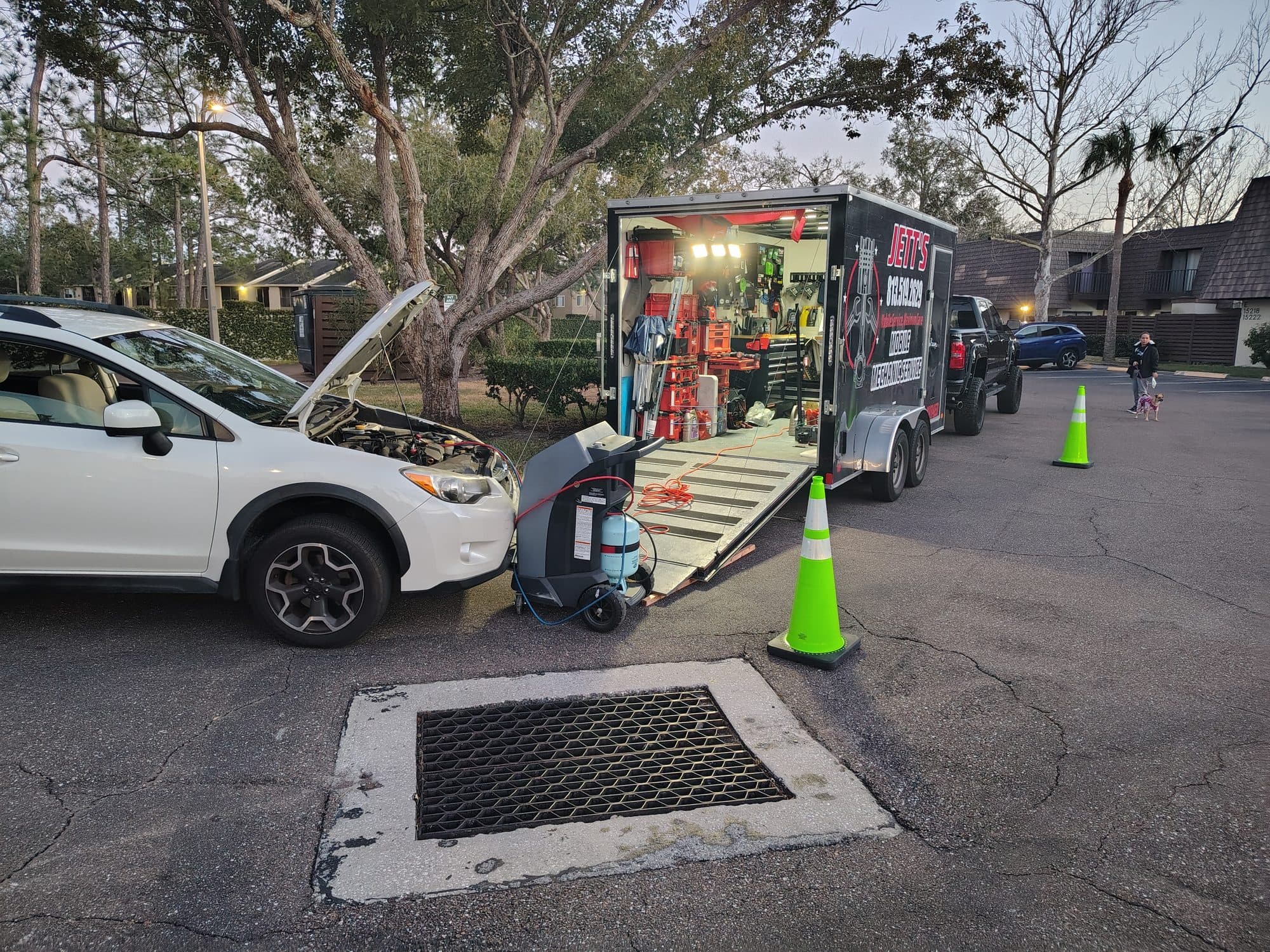 Dusk in an apartment parking lot: the trailer open and lit from inside, safety cones out, and a white Subaru with its hood up alongside