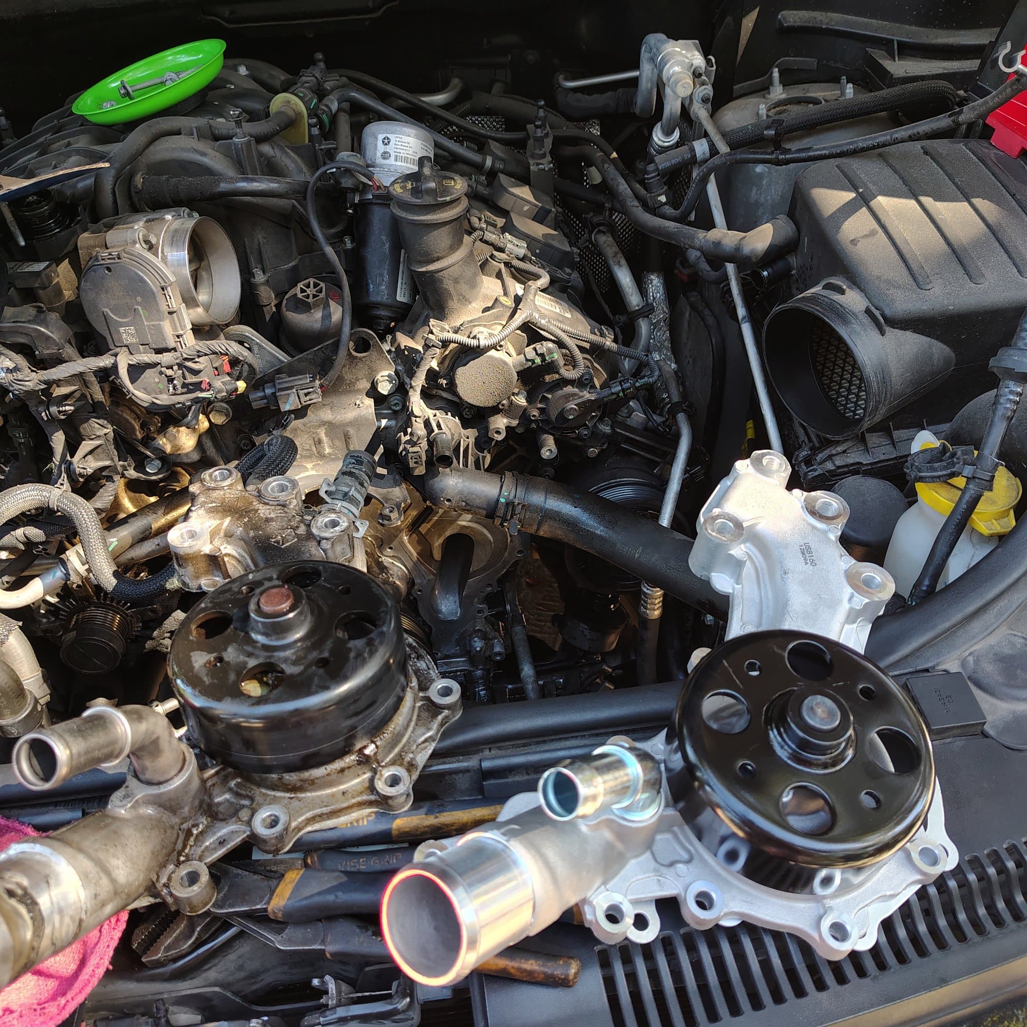 A water pump replacement: the old, grimy pump and the new one with a clean black pulley sitting side by side on the cowl above the engine, with a new thermostat housing between them